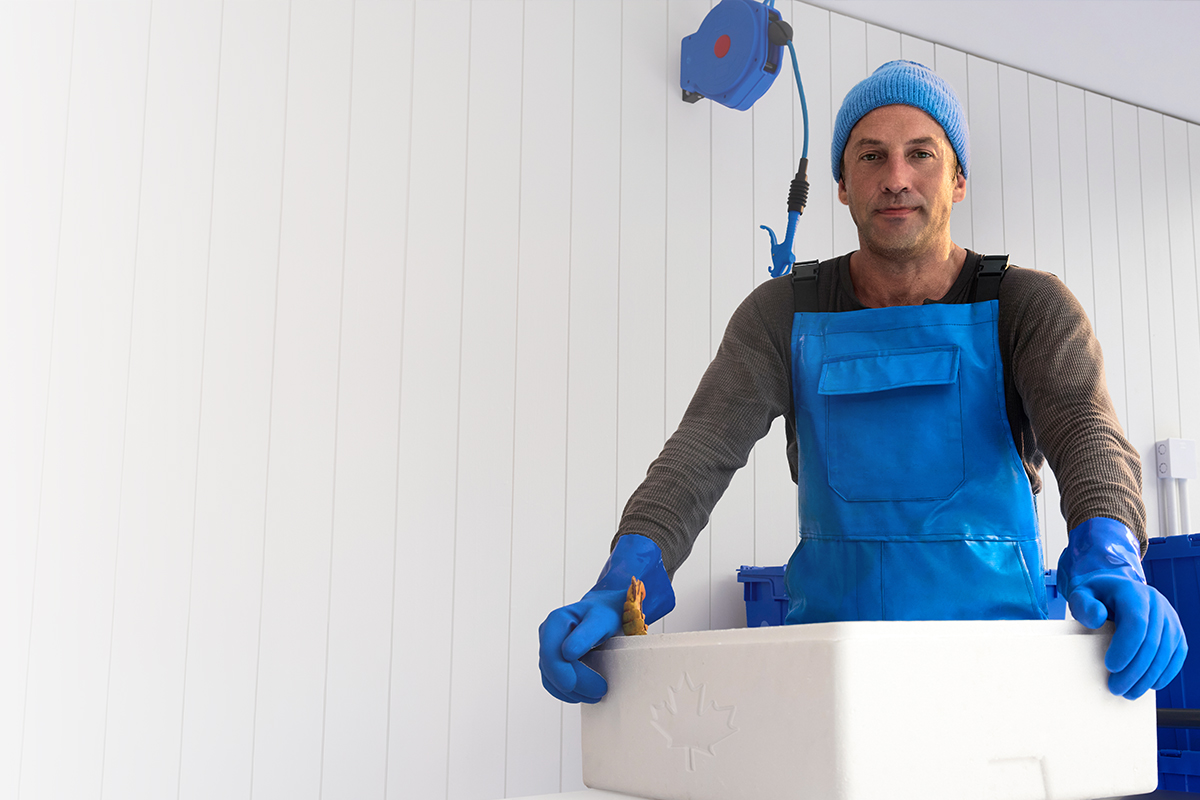 A fisherman in blue overalls and gloves is preparing to deliver seafood in blue containers with a maple leaf on them.